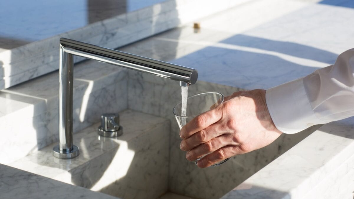 A person in a white shirt filling a glass with water from a kitchen faucet over a marble sink.